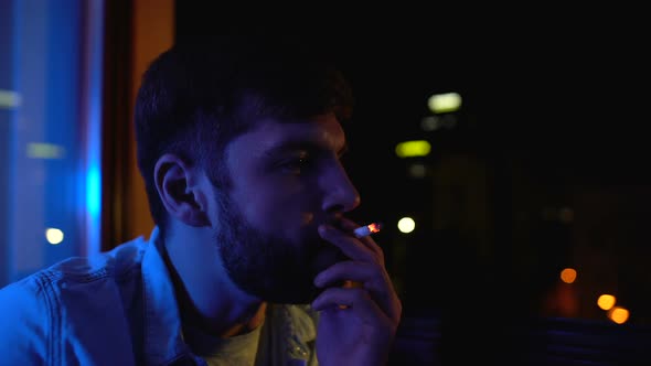 Young Man Smoking Alone Near Window at Night, Worrying About Life Problems