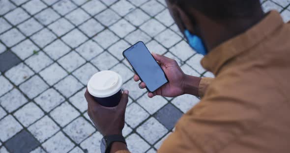 African american businessman wearing face mask using smartphone holding coffee