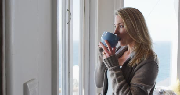 Caucasian mature woman looking through the window and drinking coffee