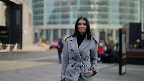 a Brunette in a Gray Coat Has Hand in Pocket and is Walking Against the Background of Modern