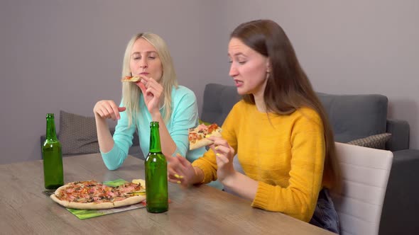 Two girls, drinking beer and eating delicious pizza, at home in their kitchen.