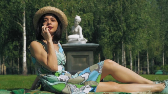 A Young Girl In a Hat Sitting On a Park Lawn And Speaks On The Phone And Smiling.