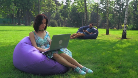 The Girl Brunette Sits In a Chair In The Park And Working On a Laptop.