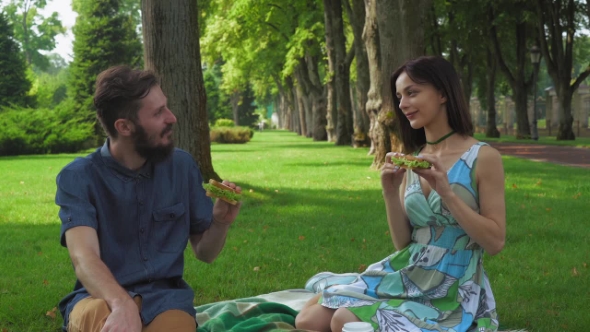 Young Couple On a Picnic In The Park Holding Sandwich.