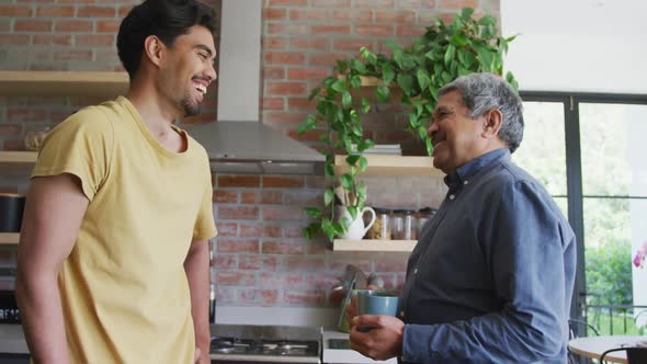Cheerful biracial father and son talking while toasting coffee mugs in kitchen at home