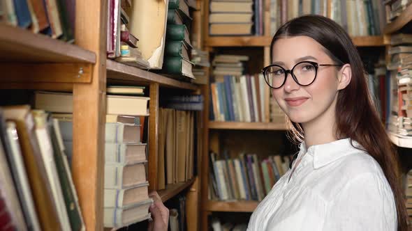 Portrait of a Beautiful Girl in Glasses Among the Book Shelves of the Library or Archive
