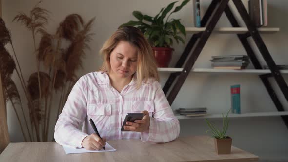 a Young Beautiful Caucasian Lady is Sitting at the Table at Home Holding Her Smartphone in Her Hands