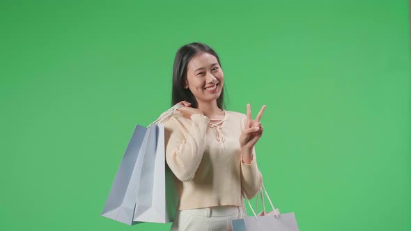 Asian Shopping Woman With Shopping Bags Making Victory Sign And Smiling In Front Of Green Screen