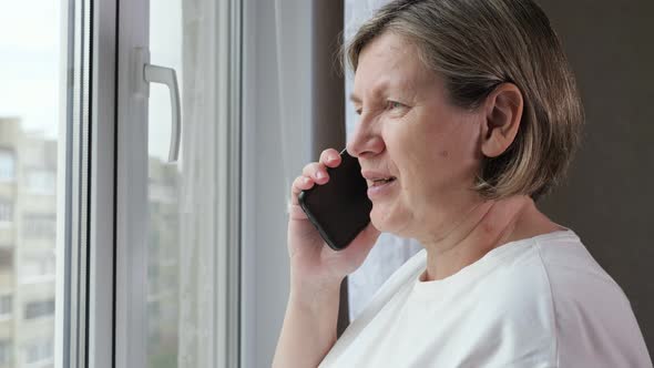 Elderly Woman in White Tshirt Talks on Phone Near Window