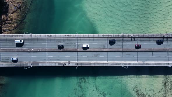 Motor vehicles and pedestrians travelling on a bridge spanning over a scenic ocean estuary. Panning