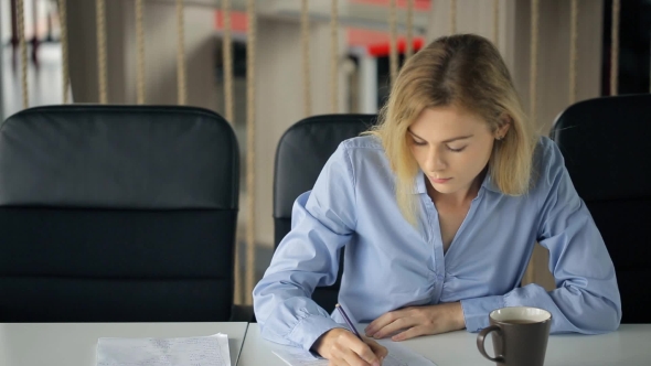 Young Attractive Blond Woman Sitting At a Table And Fills In Questionnaire With a Pencil.