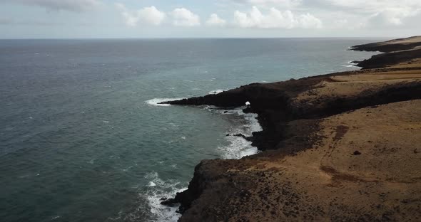 Drone shot of cliffside beach. Landscape is very brown, water is blue/grey due to clouds.