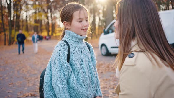 Young Mum Squatted Down and Talking to Smiling Little Daughter Schoolgirl with Backpack