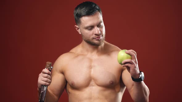 Portrait of a athletic man choosing between a fresh apple and a chocolate. Healthy food.