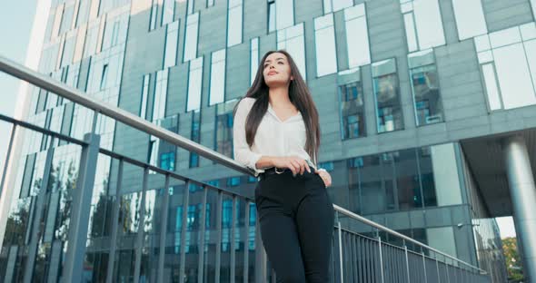Beautiful Attractive Elegantly Dressed in White Shirt Woman Stands at Railing Next to Glass Building
