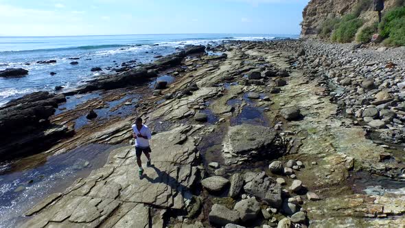 Tracking shot of a young man running on a rocky ocean beach shoreline.