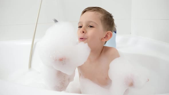 Smiling Little Boy Playing with Soap Foam in Bath and Blowing It From Hands