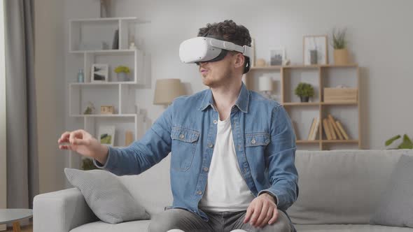 Amazed Guy Watches Online Video to VR and Moves Hands Sitting Against Window on Sofa at Home Slow