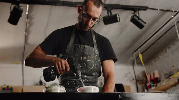 A male handyman mixes paint in a can in a workshop. A sculptor mixes resin with paint to a vase.