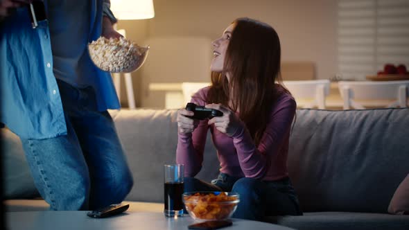 Young Woman Playing Video Game with Joystick Her Boyfriend Joining Her with Snacks and Drinks