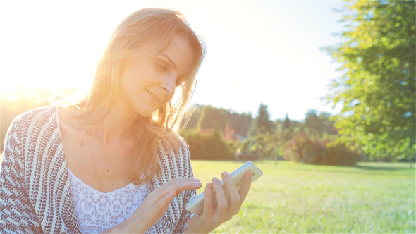 Happy Smiling Girl Using a SmartPhone in City Park Sitting on Grass 1