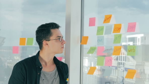 Young Handsome Man Writing On Sticky Notes On Panoramic Window In Modern Office. Brainstorming