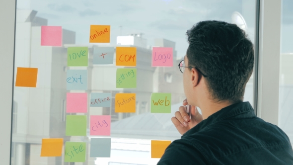 Young Handsome Man Writing On Sticky Notes On Panoramic Window In Modern Office. Brainstorming