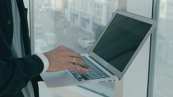 Unrecognizable Man Working On Laptop Near Panoramic Window In Modern Office. Cityscape On Background