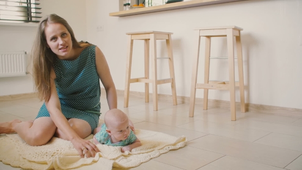 Beautiful Mother Is Helping Her Baby To Learn How To Crawl On a Blanket On The Kitchen Floor. 