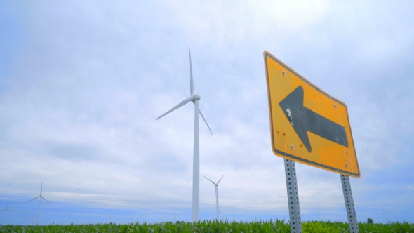 Road Sign Pointing To Wing Turbines Field Against Clouds Sky