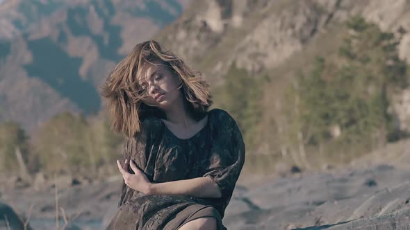 Dreaming Woman in Loose Top Dress Sits on Stone on Windy Day