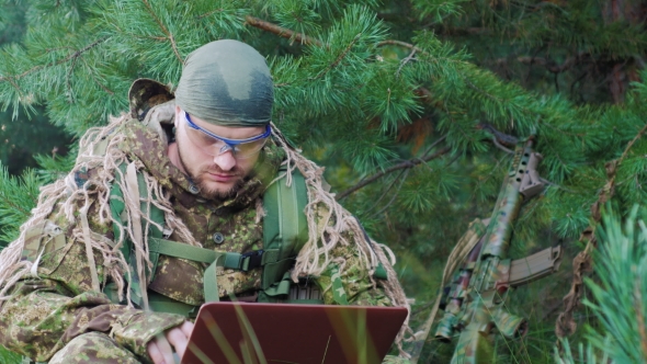 Portrait Of Military Man Sitting In The Woods, Dressed In Camouflage. It Uses Laptop alt
