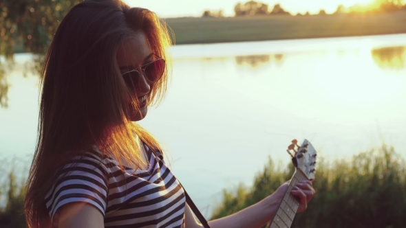 Portrait Of Young Girl Playing The Guitar Near The Evening Lake. 