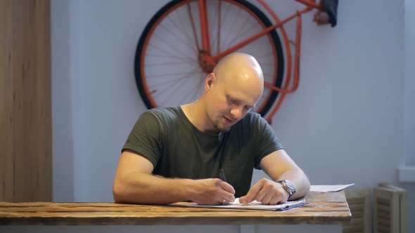 Bald Bearded Man Sits At Wooden Table In Studio.