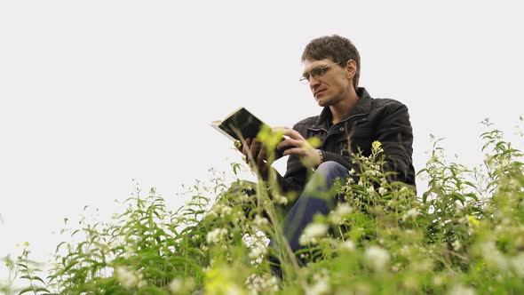 Man Reading Book in Green Hilly Field. Side View of Young Handsome Man in Glasses and Warm Jacket