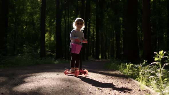 Little Girl Kid on Orange Scooter Stands on Asphalt Road in Forest Park