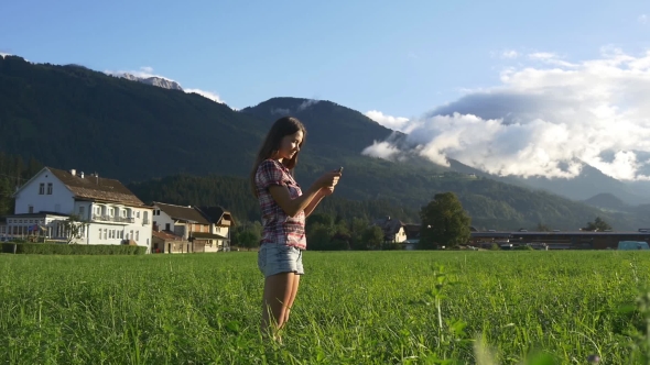Woman Texting On Phone In Green Field At Mountain