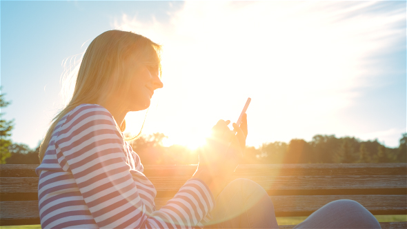 Happy Smiling Girl Using a Smartphone in a City Park Sitting on a Bench 