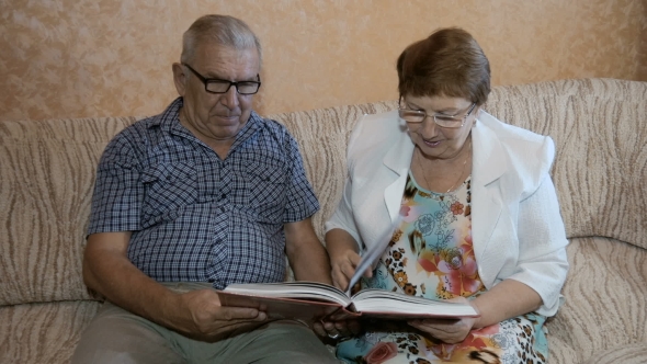 Beautiful Couple In Age, Reading a Book Sitting On The Couch At Home.