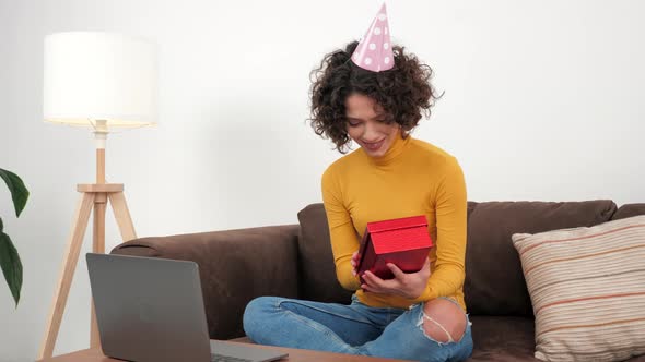 Happy Woman in Party Hat Opens Gift Box and Surprised Sitting in Couch at Home