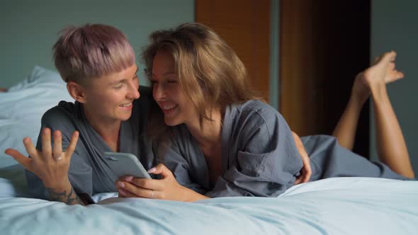 Cheerful Young Lesbian Couple Laughing While Using Their Smartphones in the Bed