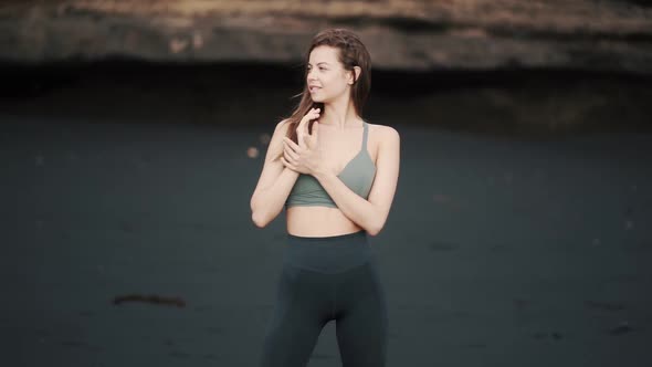 Young Sexy Woman in Sportswear Poses on Black Sand Beach Looks at Camera Smiles