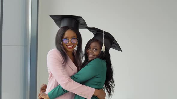 Two Girlfriends Graduates in Festive Costumes and Master's Hats Hug and Look at the Camera with a
