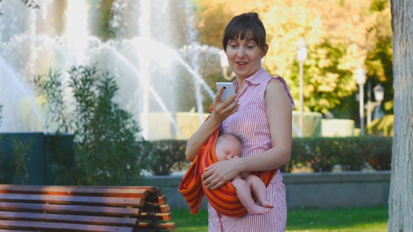 Young Woman With Sleeping Baby in Sling Happily Surprises About Phone Message