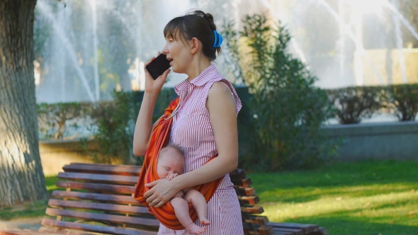 Young Woman With Sleeping Baby in Sling Starts Phone Call