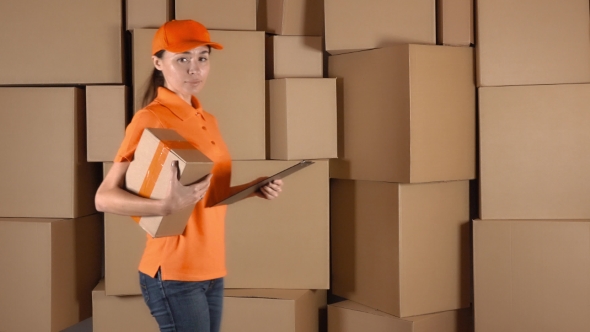Beautiful Female Courier In Orange Uniform Delivering a Parcel Against Carton Stacks Backround