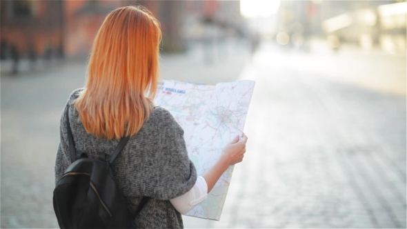 The Young Red-haired Female Tourist Attentively Considers The Map. The Girl Walks Streets