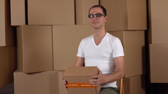 Young Geeky Owner Of Small Internet Store Holding Parcel Against Carton Stacks.  Clip