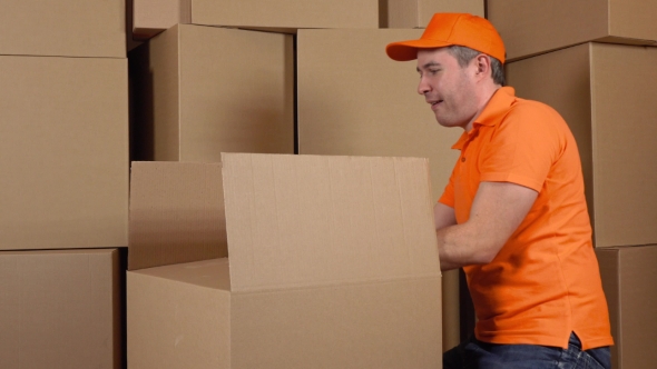Warehouse Worker In Orange Uniform Packaging Big Carton With Duct Tape Gun. Multiple Boxes