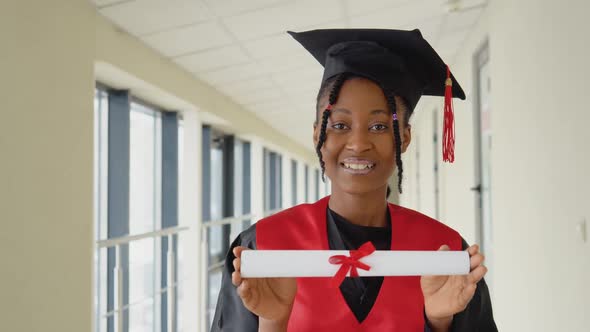 African American Female Graduate in Mantle Stands with a Diploma in Her Hands and Smiles